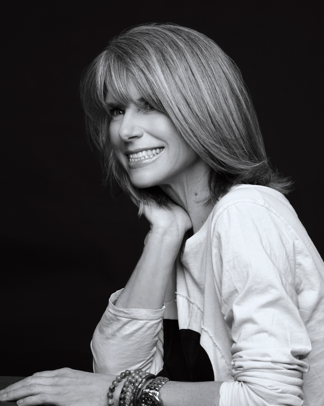 Black and white professional portrait of Judy Brooks smiling warmly, resting her hand near her chin, wearing a light long-sleeve shirt and bracelet against a dark background.