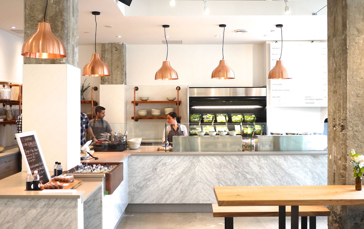 right modern café interior with marble counters, copper pendant lights, and staff preparing food — representing Field & Social’s clean, intentional approach to nourishment and community-focused dining.