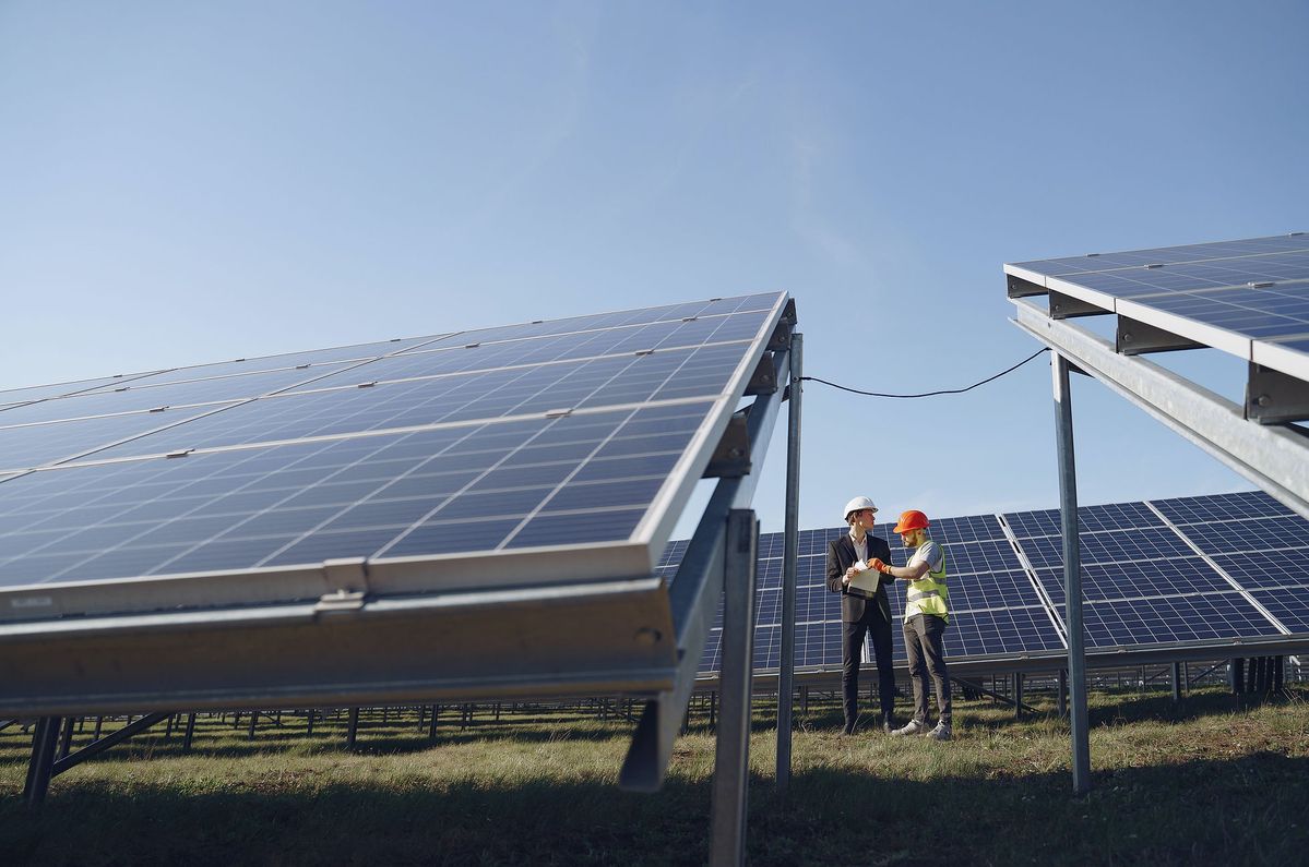 Two engineers wearing safety helmets and vests inspect solar panels at a large solar energy farm under a clear blue sky, representing renewable energy and sustainability.