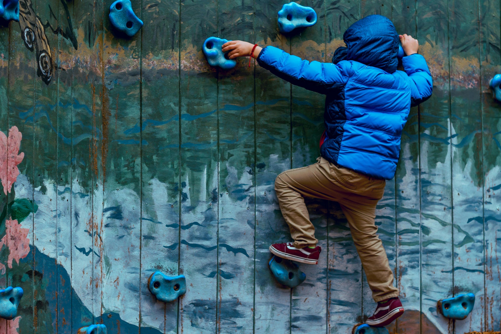 Child climbing a colorful wall with blue holds — symbolizing Brainchild’s focus on growth, persistence, and learning through creative challenge and exploration.