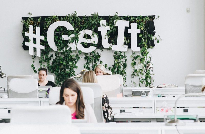 Modern open office space with people working at desks in front of a wall decorated with plants and large white letters spelling "#GetIt".